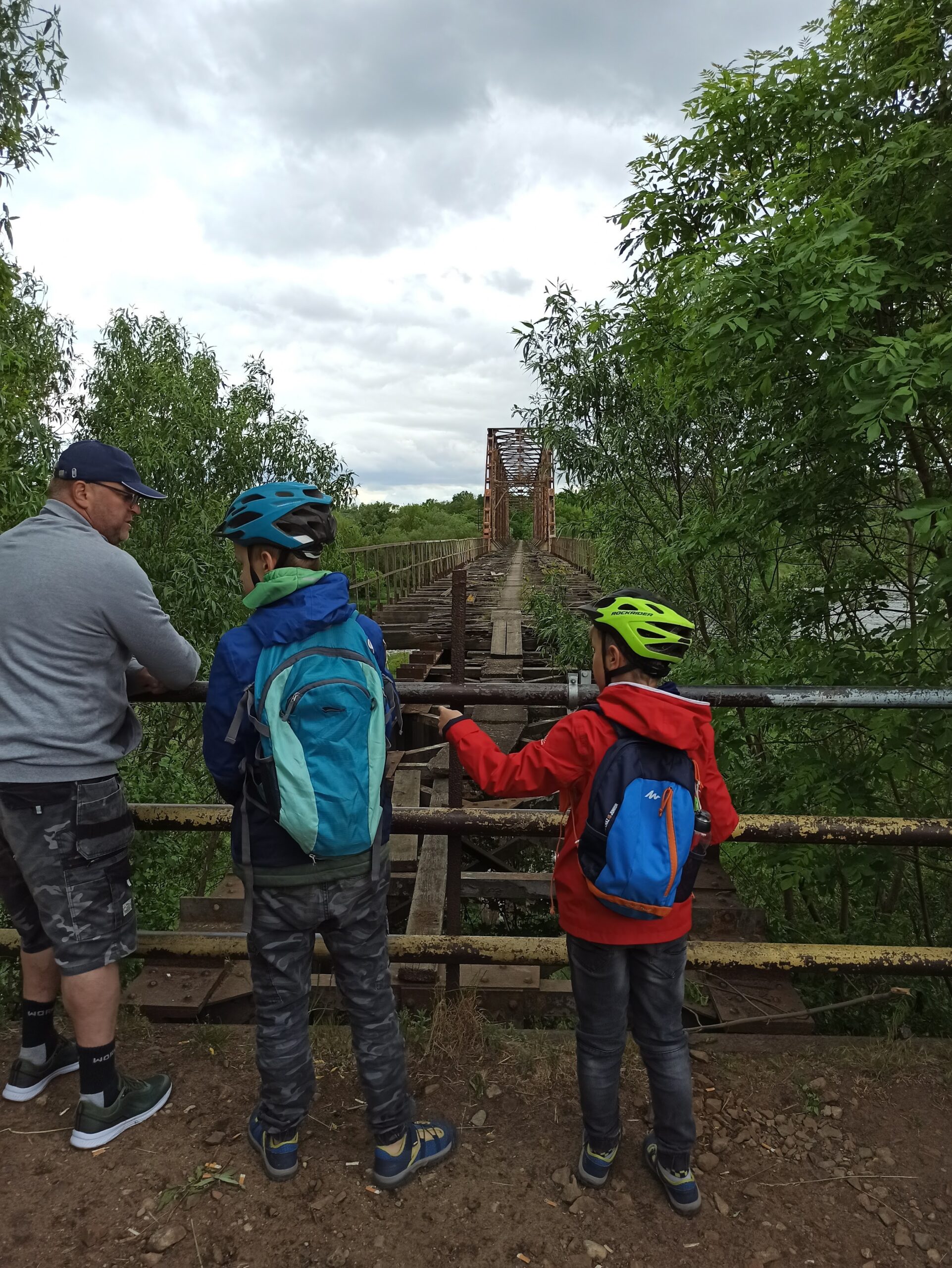 Na zdjęciu widoczne trzy osoby. Mężczyzna i dwóch chłopców. Stoją przy barierach przed mostem kolejowym.