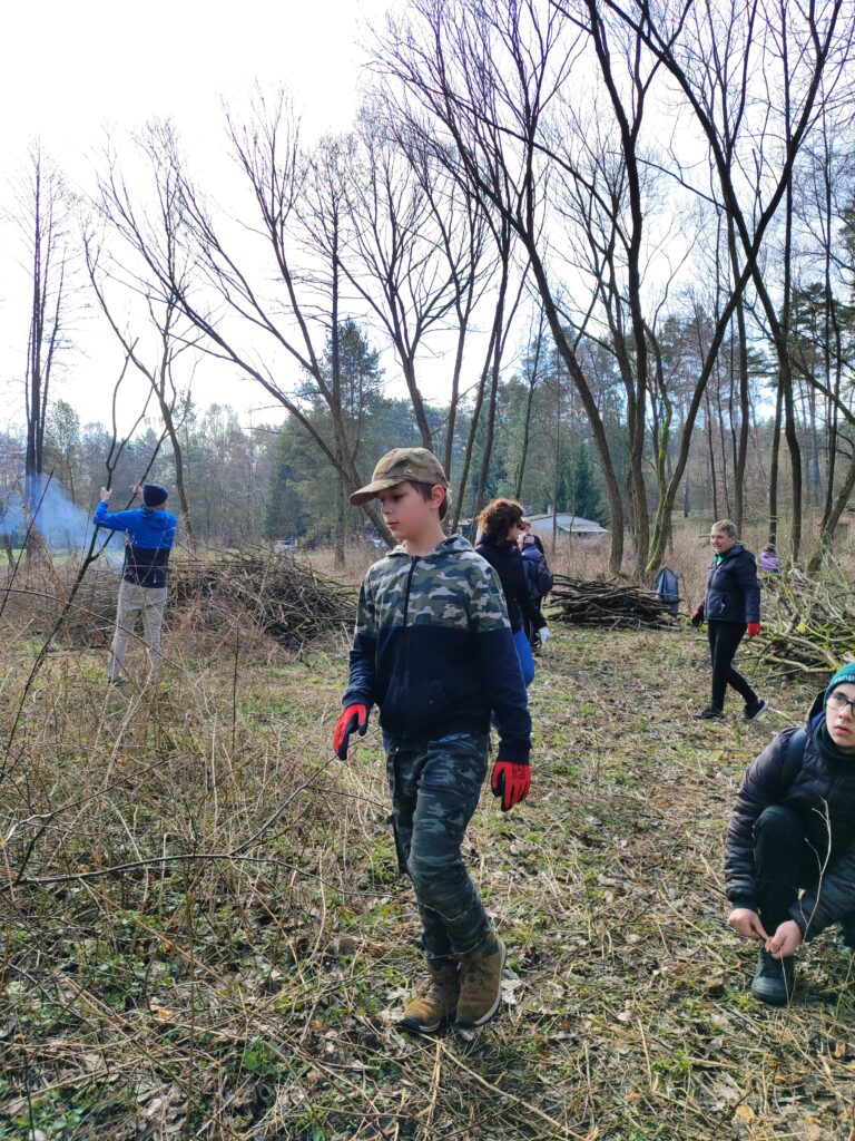 nastolatek w czapce i czerwono- czarnych rękawicach podczas akcji sprzątania lasu. w tle inne osoby i dym z ogniska